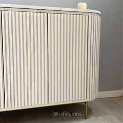 Mueble de madera con diseño estriado en color blanco, con patas doradas y una vela encendida sobre la parte superior.