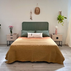 Una cama decorada con un cabecero verde, almohadas grises y una manta amarilla. A los lados, hay mesas de noche con flores y una lámpara decorativa.