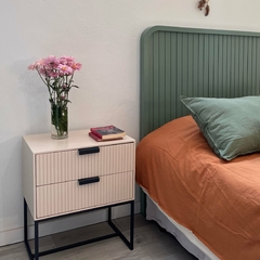 Cama con cabecera verde, almohada verde, sábanas naranjas y mesa de luz con flores y libros.