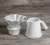 Kit de Jarra e Coador de Café V60-02 em Cerâmica com esmaltação branca da Felline Cerâmica. O Coador está encaixado no corta pingos e ao lado a jarra também na cor branca. O conjunto está parte de perfil sobre uma mesa rústica de madeira.
