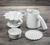 Kit de Jarra e Coador de Café V60-02 em Cerâmica com esmaltação branca da Felline Cerâmica. Vista de 4 peças deste conjunto sobre uma mesa de madeira, coador, jarra, prato de apoio e corta pingos. 