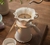 Chaleira derramando água quente sobre pó de café no filtro de papel que está encaixado na boca de uma jarra. O Kit de Jarra e Coador de Café V60-02 é de cerâmica esmaltado em branco, feito à mão pela Felline Cerâmica e está sobre uma mesa de madeira.  