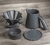 Kit de Jarra e Coador de Café V60-2 em Cerâmica com esmaltação preta da Felline Cerâmica. Vista de 4 peças deste conjunto sobre uma mesa de madeira, coador, jarra, prato de apoio e corta pingos. 