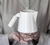 Vista de perfil de uma jarra de cerâmica para café e leite apoiada sobre duas mãos feminina em frente do corpo. O conjunto é esmaltado em branco semifosco. A peça é feita à mão pela Felline Cerâmica.