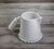 Vista de perfil de uma jarra de cerâmica para café e leite apoiada sobre sobre uma mesa de madeira. O conjunto é esmaltado em branco semifosco. A peça é feita à mão pela Felline Cerâmica.
