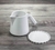 Vista de perfil de uma Jarra de cerâmica para café e leite com prato de apoio. Este ângulo valoriza o pegador da jarra. O conjunto é esmaltado em branco semifosco. Estão sobre uma mesa de madeira rústica. Este ângulo valoriza o bico da jarra.
