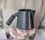 Vista de perfil de uma jarra de cerâmica para café e leite apoiada sobre duas mãos feminina em frente do corpo. O conjunto é esmaltado em preto semifosco. A peça é feita à mão pela Felline Cerâmica.