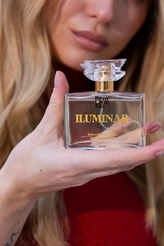 A imagem mostra uma mulher segurando um frasco de perfume com as duas mãos. O frasco tem a etiqueta "ILUMINAR" em letras douradas e informações adicionais do produto. A mulher tem cabelos loiros, esmalte claro nas unhas e está parcialmente visível, com de