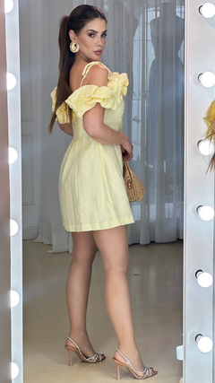 Modelo posando com vestido curto amarelo de linho, com babados nas mangas, segurando uma bolsa enquanto olha para a câmera.