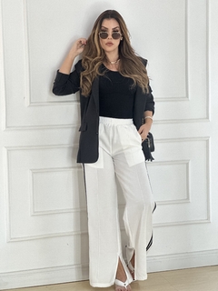 Mulher usando calça branca de viscose com listras pretas, blusa preta e blazer, posando em frente a uma parede branca.