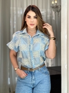 Mulher posando com uma camisa azul clara bordada de manga curta e calça jeans, em ambiente iluminado.