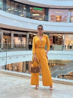 Modelo vestindo um conjunto de linho amarelo, composto por blazer curto e calça ampla, posando em um shopping.