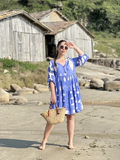 Mulher usando um vestido curto azul com estampa, segurando uma bolsa de palha, em uma praia com casas ao fundo.