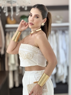 Modelo vestindo um conjunto Caribe com top branco e calça, acessórios dourados e maquiagem elegante, em um ambiente claro.