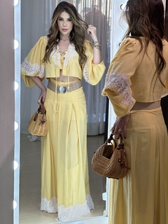 Model usando um conjunto amarelo com detalhes em renda, segurando uma bolsa de palha e posando em frente ao espelho.