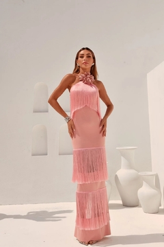 Modelo usando um vestido longo rosa com franjas, destacando um detalhe de flor no pescoço, em um cenário minimalista.