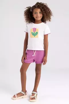 Menina usando um conjunto infantil de meia malha e moletom da Bugbee, com blusa branca e estampada de flor e shorts roxos.