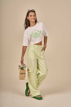 Modelo usando blusa juvenil cropped branca com estampa de borboleta verde e calças largas amarelo-claro, segurando um vaso de flores.