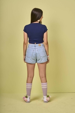 Modelo feminina de costas usando shorts saia juvenil jeans com pregas, em um fundo amarelo.