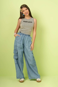 Modelo vestindo calça juvenil wide leg utilitário em chambray denim, com blusa cinza e acessórios coloridos, em fundo verde.