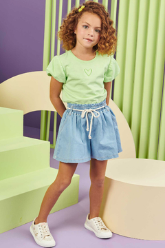 Menina usando shorts infantil de jeans claro com elástico e camiseta verde em ambiente colorido.