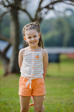 Menina sorridente usando um conjunto de blusa listrada e shorts laranja, com flores no cabelo e fundo natural.