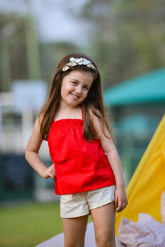 Menina sorrindo vestindo um conjunto de blusa vermelha e shorts claros, em um ambiente ao ar livre.