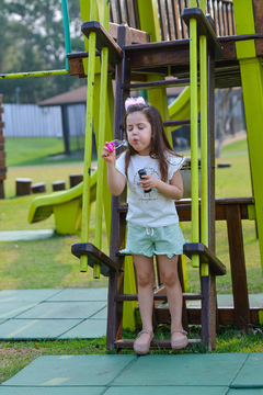 Criança brincando em um parque, usando um conjunto de blusa e shorts na cor verde claro, soprando bolhas de sabão.