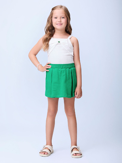 Menina sorrindo usando uma regata branca e shorts verdes, posando com as mãos na cintura.