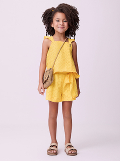 Menina sorrindo usando um conjunto amarelo de laise com laço, composto por blusa e short, com uma bolsa pequena.