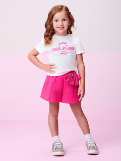 Menina sorridente usando um conjunto rosa com laço de strass, camiseta branca com estampa colorida e shorts rosa.