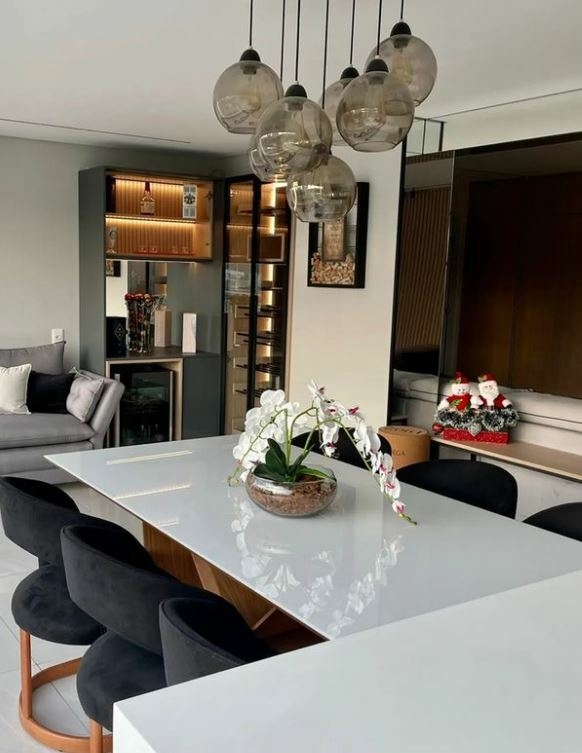 Imagem de uma sala de jantar moderna com luminárias pendentes de vidro fumê, mesa branca e cadeiras pretas, decorada com uma planta de orquídea.
