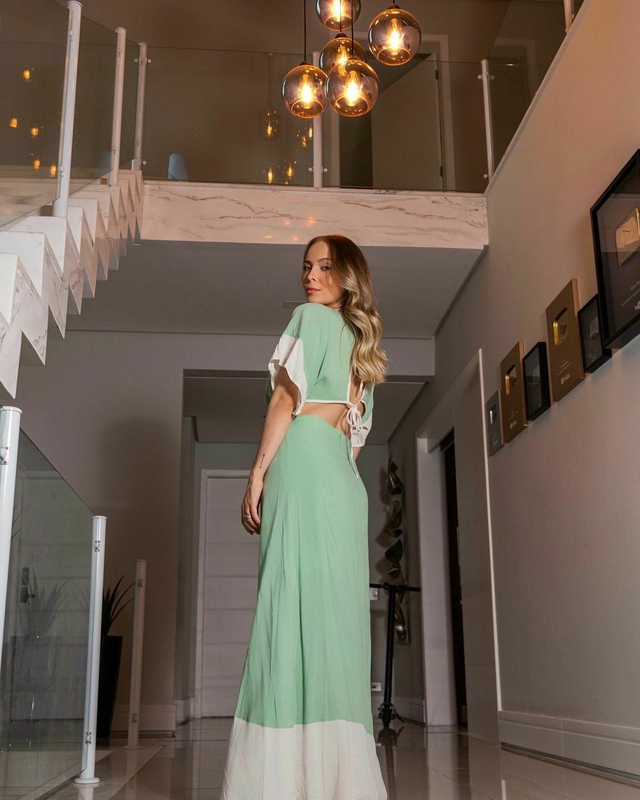 Tata Cocielo usando vestido longo verde com detalhes brancos em um ambiente moderno, com lustres pendentes ao fundo.