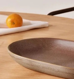 Centro de mesa minimalista em cerâmica rosa nude com formato oval, posicionado sobre uma mesa de madeira, ao lado de uma laranja e um pano branco.