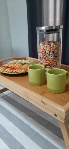 Bandeja de bambú con patas, decorada con tazas verdes y un plato de frutas y galletas, acompañada de un frasco de cereales de colores.