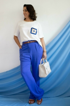 Modelo vestindo blusa branca de manga curta com detalhe em crochê azul, calça azul, posando em fundo azul.