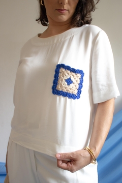 Blusa de manga curta na cor branca com detalhe em crochê azul e bege no bolso.