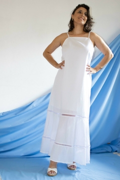 Modelo exibindo um vestido longo branco, com alças finas e detalhes em renda, contra um fundo azul suave.