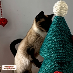 Gato se esfregando em uma árvore de Natal arranhador verde, decorada com enfeites vermelhos e uma estrela branca no topo.