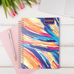 Planner BIG abstrata da Coleção Dandelion 25 com capas coloridas e vibrantes, sobre uma mesa com flores e um caderno rosa.