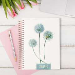 Planner A5 da Coleção Dandelion com flores azuladas, ao lado de um caderno rosa e uma caneta.