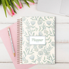 Planner A5 da coleção Dandelion com capa floral, em cima de uma mesa com um caderno rosa e uma caneta.