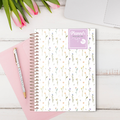 Planner BIG flores delicadas da Coleção Dandelion 25 sobre uma mesa com flores e um caderno rosa ao lado.