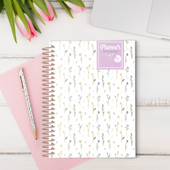 Planner A5 com estampa de flores delicadas da Coleção Dandelion 25, ao lado de uma caneta e um caderno rosa.