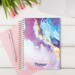 Planner grande com design de mármore colorido, coleção Dandelion 25, sobre uma mesa com flores e caneta.