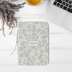 Caderno pocket da coleção Dandelion com estampa de flores e folhas, disposto sobre uma mesa com um copo de chá e um laptop ao fundo.