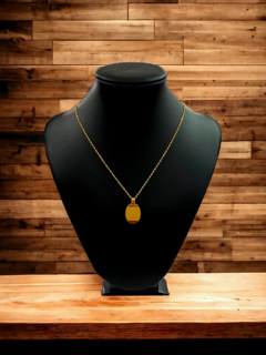 Collar dorado con un colgante ovalado sobre un maniquí negro, frente a un fondo de madera