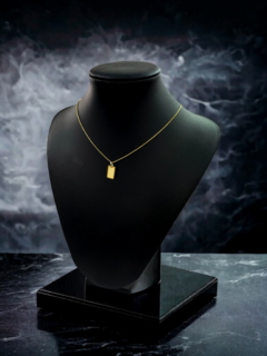 Collar de oro con un colgante rectangular, exhibido en un soporte negro sobre fondo nebuloso.