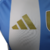 A nova camisa titular da Argentina destaca o branco com três listras verticais azul celeste. Detalhes azuis estão presentes nas mangas, gola. Logos Adidas e AFA em dourado e o patch de campeã mundial no peito.