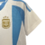 A nova camisa titular feminina da Argentina 2024 destaca o branco com três listras verticais azul celeste. Detalhes azuis estão presentes nas mangas, gola. Logos Adidas e AFA em dourado e o patch de campeã mundial no peito.